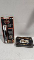 Photo showing black flashlight with silver top and black leather holder in original packaging, Harley Davidson branded collectible tin with motorcycle graphic.