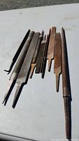 Photo showing a collection of 8 metal files on a light surface, arranged side by side, displaying varying lengths and shapes including flat and round files with signs of rust and wear.