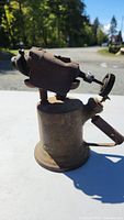 Side view of vintage metal torch showing brass base, side handle, and rusted burner top with valve knob