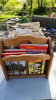 Wooden magazine holder filled with assorted vintage magazines and flyers including a British Columbia travel brochure and other colorful papers.