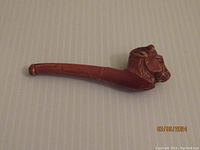 Child's clay bubble pipe photographed from one side, showing the long reddish-brown curved pipe with a camel head sculpted at the bowl end.