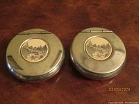 Closed view of two round English pewter hinged boxes with engraved medallions on lids.