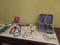 Group photo showing eleven costume necklaces laid out on a dark wooden surface including a decorative blue box on the right holding two smaller necklaces. Multiple strands of beads in white, purple, turquoise, and other colors are visible, along with a large silver-tone pendant