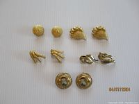 Wide view showing all five pairs of gold tone Monet clip-on earrings arranged in two rows on plain white background.