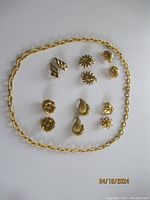 Top view of six pairs of Monet gold-tone clip-on earrings arranged in two rows with a gold-tone chain necklace surrounding them laid flat on white background.