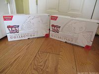 Two boxes of Pokémon Scarlet & Violet 151 Ultra-Premium Collection placed side by side on wooden floor, showing front of both boxes and Mew artwork.
