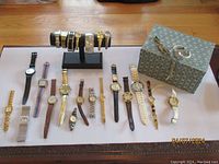 Overhead photo showing over 20 wristwatches laid out on a white surface including assorted metal and leather strap watches and a decorative box.