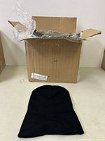Open cardboard box containing the black knit toque hats with one hat displayed outside the box showing its shape and texture.