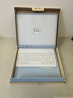 Open cardboard box showing white wireless keyboard inside a blue case, keyboard wrapped in plastic, indicating unused condition.