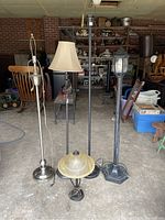 View of five lamps grouped together showing one silver tall lamp with rusted base, two tall black lamps including one with beige shade, one indoor table lamp with glass shade, and one tall outdoor lamp post with lantern top.