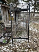 Large black metal bird cage standing outdoors on grass and snow, showing front and top section. The cage has a rectangular bottom tray and thin metal legs.