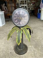 Front view of double-sided clock stand resembling a palm tree with green metal leaves and round clock face with Roman numerals.