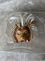 Top-down view of the Disney Lion King souvenir mask inside a clear plastic container showing the lion face and feathered mane.