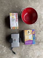Top-down view of all four items: red metal colander, boxed Starfrit fry cutter, boxed nesting storage containers, and waffle iron unit closed with power cord.