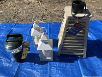 Photo of four lanterns arranged on blue tarp outdoors: one green metal camp lantern, two white plastic candle lanterns, one tall wooden slatted candle lantern with black top and tag