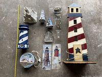 Top-down view of all items in the lot showing arrangement, including the largest wooden lighthouse light, ceramic lighthouses, ships in bottles, metal wall art, glass paperweight, and plastic lighthouse.