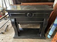 Front view of black wooden mantle cover with decorative molding and central circular design, showing surface wear.