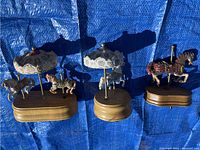 Front view of three wooden horse music boxes, two carousel style with horses spinning beneath decorated canopies, one single horse figure with mechanical parts on wooden base.
