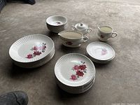 White ceramic plates and bowls set with ribbed edges and a rose pattern in pink and red tones, displayed in groups by size, along with matching gravy boat, sugar bowl with lid, and cream jug on a concrete floor.