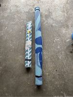 Two rolled outdoor rugs, one smaller teal and white, one larger blue and gray.