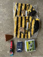 Two unused bee dog costumes in plastic packaging, red dog collar, and boxed training devices placed on concrete floor.