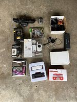 Top view of all electronic items arranged on floor including boxed Coca-Cola backup radio, boxed electronics, gaming discs, and various wires/cameras.