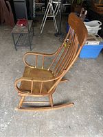 Side view of wooden rocking chair showing slatted backrest, curved armrest, and rocker base with visible wear and damage.