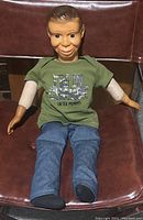 Full body image of the 26-inch ventriloquist dummy seated on a brown chair, showing outfit and physical condition
