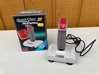 Photo of QuickShot XII joystick controller, gray with red button, placed beside original box showing product branding and compatibility text.