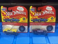 Pair of Hot Wheels cars in original blister card packaging. Yellow and maroon cars with matching blue buttons. Cards feature Hot Wheels Vintage Collection branding and flame graphics.
