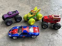 Four toy vehicles displayed: a blue and red Superman Ford GT diecast car with large chrome wheels and three monster trucks in purple, green, and red colors.