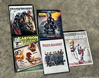 Five DVDs/Blu-rays arranged on concrete surface, showing Transformers, Star Wars Rogue One, Cartoon Classics, Police Academy, and Robotech covers.