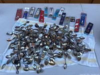 Wide view of a large collection of over 100 vintage souvenir spoons laid out on a towel, showing a mix of metal and enamel spoons with various handle designs and some boxed spoons.