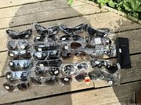 Top-down view of 24 new old stock sunglasses, all individually wrapped, showing a wide variety of frame and lens styles/colors spread on wooden surface.