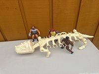 Three MOTU figures standing on a bone-colored dinosaur skeleton carrying case, displayed against a wooden background.