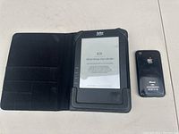 Photo of Kobo eReader in black folio case next to older Apple iPhone with black plastic back, placed on a light surface.