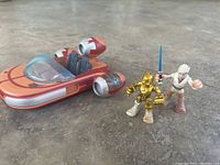 Red and silver land speeder toy with C-3PO and Ben Kenobi figures standing beside it on a floor