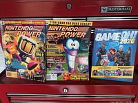 Two Nintendo Power magazines and one Game On 2020 gaming book on a red surface.