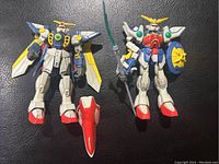 Two vintage Gundam action figures with one holding a green spear and round shield, the other with large wing attachments and a red accessory between their feet.