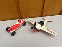 Red and white toy race car and white toy airplane photographed side by side on a flat surface in front of a wood-paneled wall.