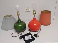 Pair of retro lamps with mismatched shades, one green base and one orange base, both with light bulbs and two-prong plugs visible.