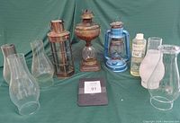 Full view of five antique oil lamps with varying styles, five spare glass chimneys and a bottle of lamp oil on green fabric background