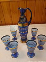 Blue glass decanter surrounded by six matching blue glasses, all with gold and black stripes and gold leaf design on decanter.