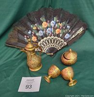 Small brass teaset comprising a 6 inch pot, two 3 inch cups, a lidded bowl, and a decorative floral fan, shown against a green backdrop.