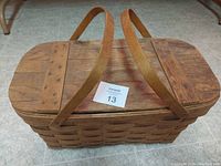 Classic woven wicker picnic basket with wooden slats on top and two carrying straps lying on gray tile floor