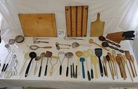 Photo showing two large wooden cutting boards, various kitchen utensils including wooden spoons, metal whisks, ladles, spatulas, strainers, tongs, and knife block with knives.