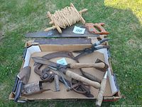 Photo of the full lot showing various rusty old hand tools, a large saw, garden shears, a rope spool, trowel, cultivator, and pipe wrench inside a cardboard box outdoors.