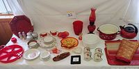 Wide view of all red and white kitchen and stoneware items on white table including plates, cups, trays, vases and towels.
