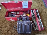 Open red metal toolbox displaying assorted vintage hand tools inside including clamps, wooden mallet, and leather tool belt