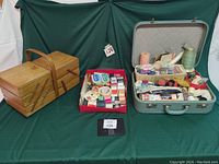 View of full lot showing a wooden accordion box, a red box with sewing threads, and a retro vintage suitcase opened displaying sewing supplies and notions.